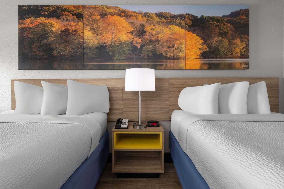 A hotel room with two beds, a nightstand with a lamp, and a large autumn-themed painting above the beds.