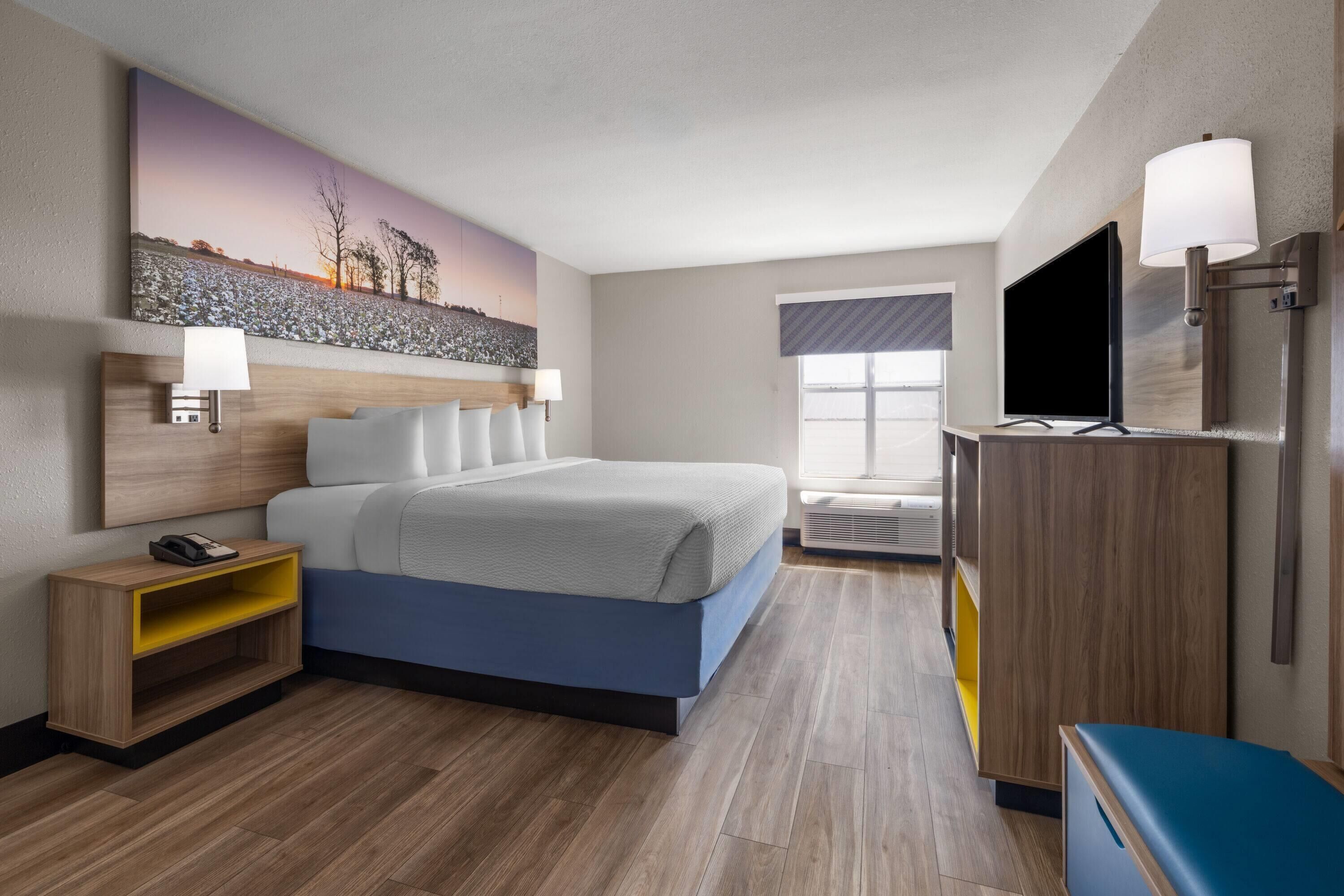 A modern hotel room with a bed, bedside table, lamp, TV, and a painting on the wall.