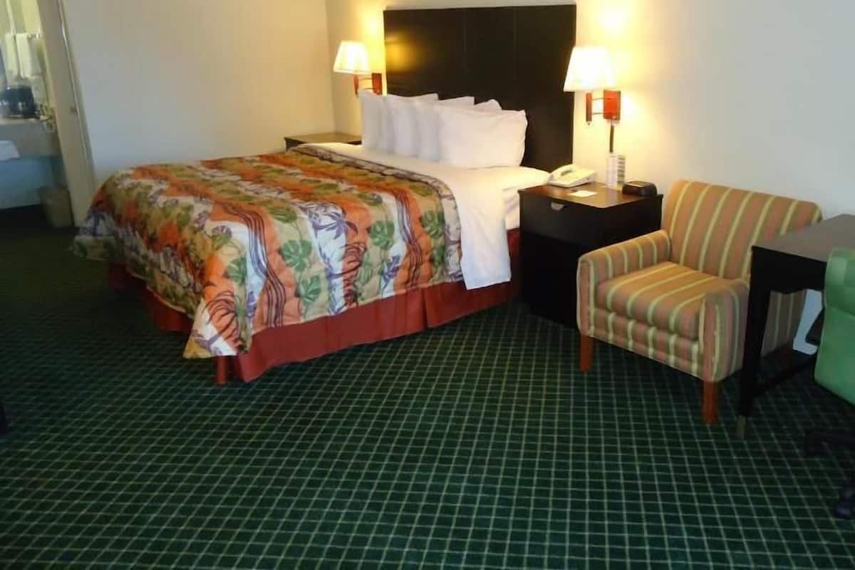 A hotel room with a bed, two bedside lamps, a chair, a small table, and a green checkered floor.