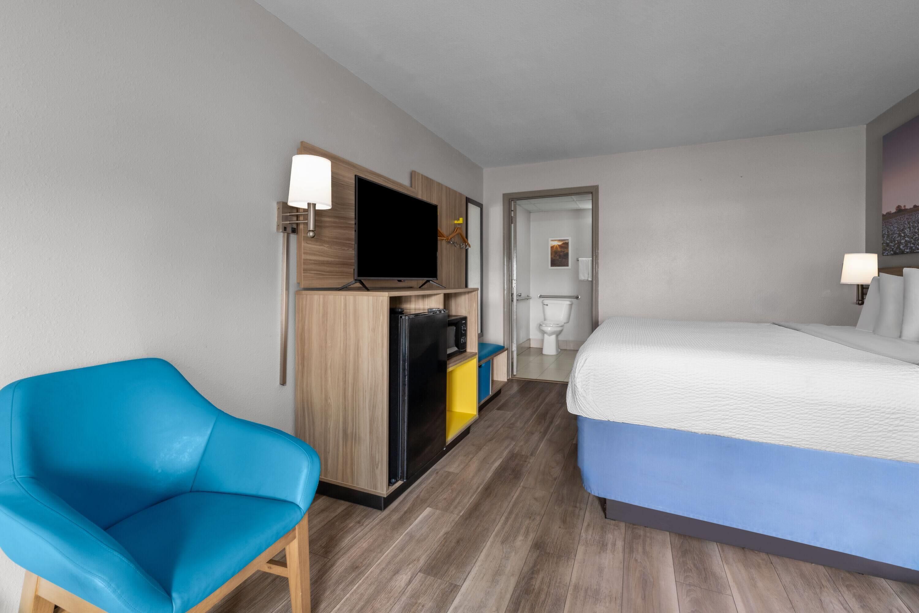 A hotel room with a bed, a television, a minibar, a blue chair, and a bathroom visible through an open door.