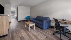 A modern, well-lit room with a wooden floor, a blue sofa, a small wooden table, a blue chair, and a black office chair. - Days Inn by Wyndham Alma (Alma)