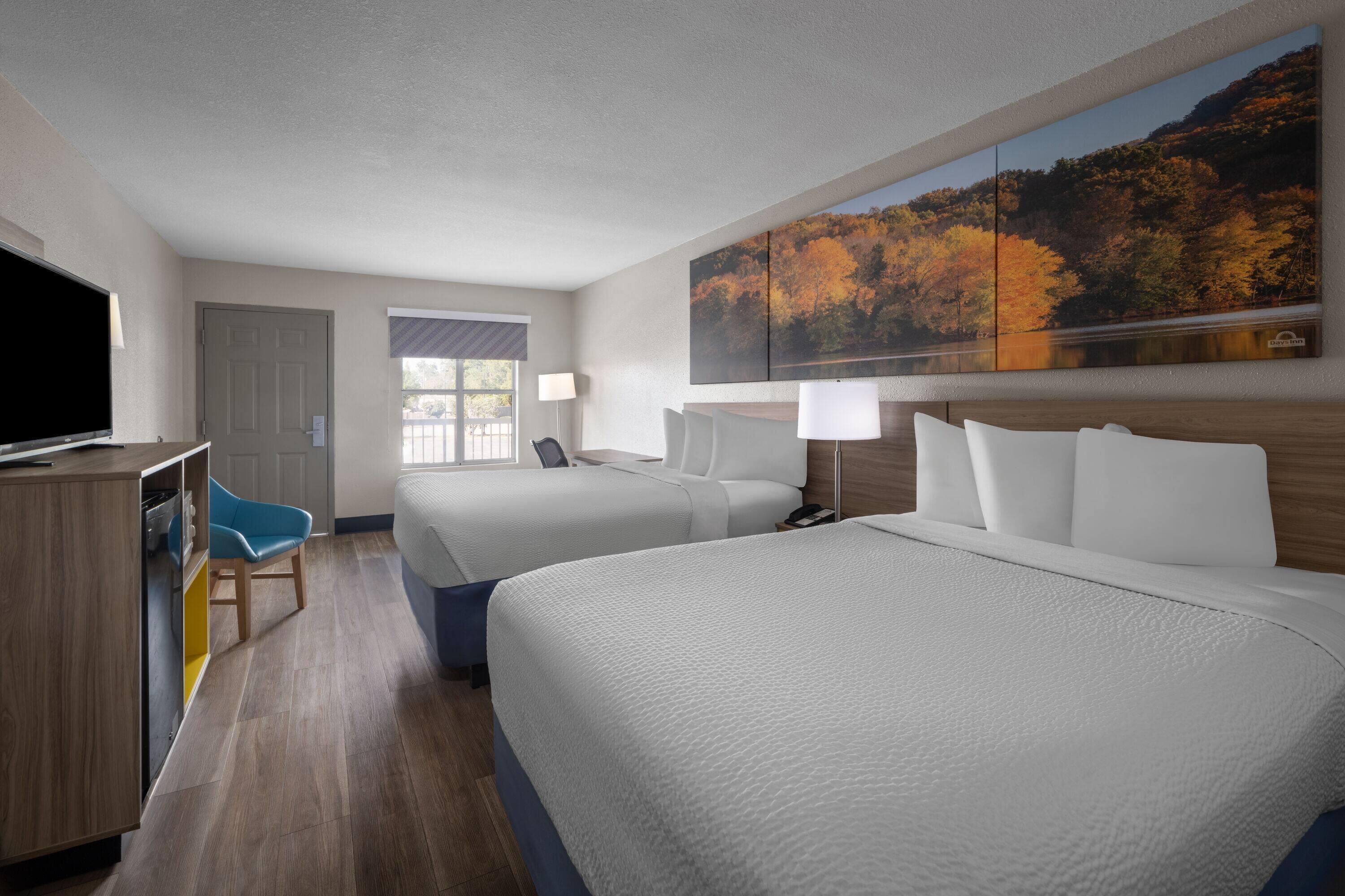 A hotel room with two beds, a desk with a TV, a chair, and a large painting of autumn foliage.