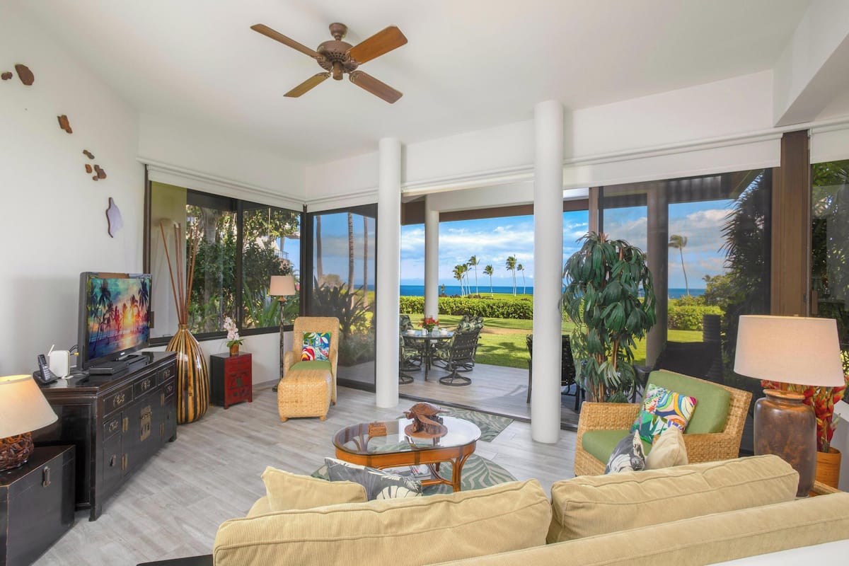 A spacious living area with a large sofa, a coffee table, and a television. There is a ceiling fan, a large window with a view of the ocean, and several pieces of furniture including a chair and a cabinet.