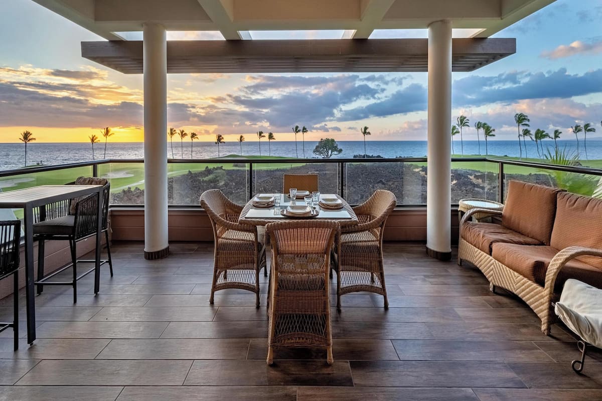 A covered outdoor seating area with a table and chairs, overlooking a golf course and ocean at sunset.