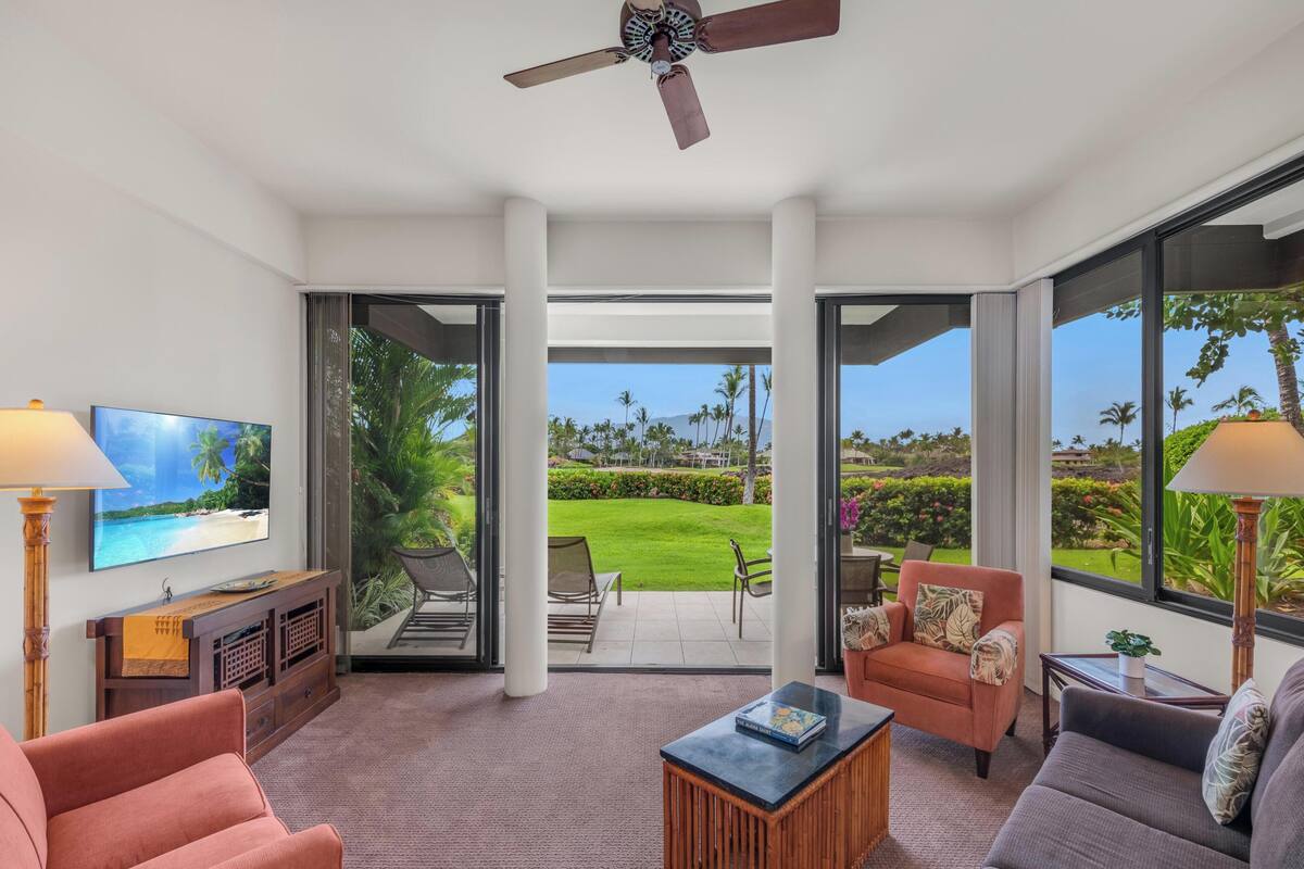 A spacious living area with a TV, comfortable seating, a ceiling fan, and large windows offering a view of a lush garden and beach.