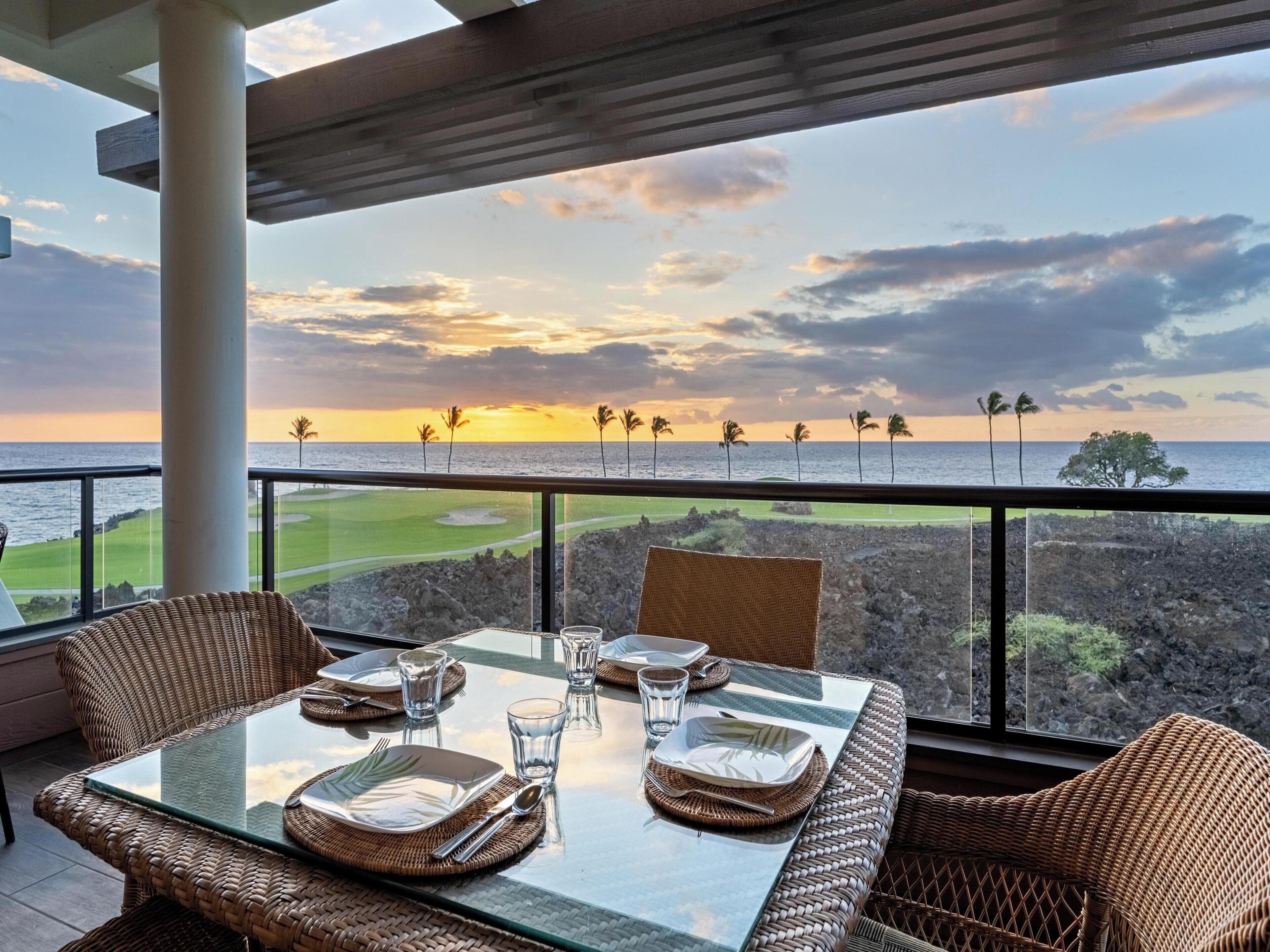 A dining table set for two with a view of the ocean and palm trees at sunset.