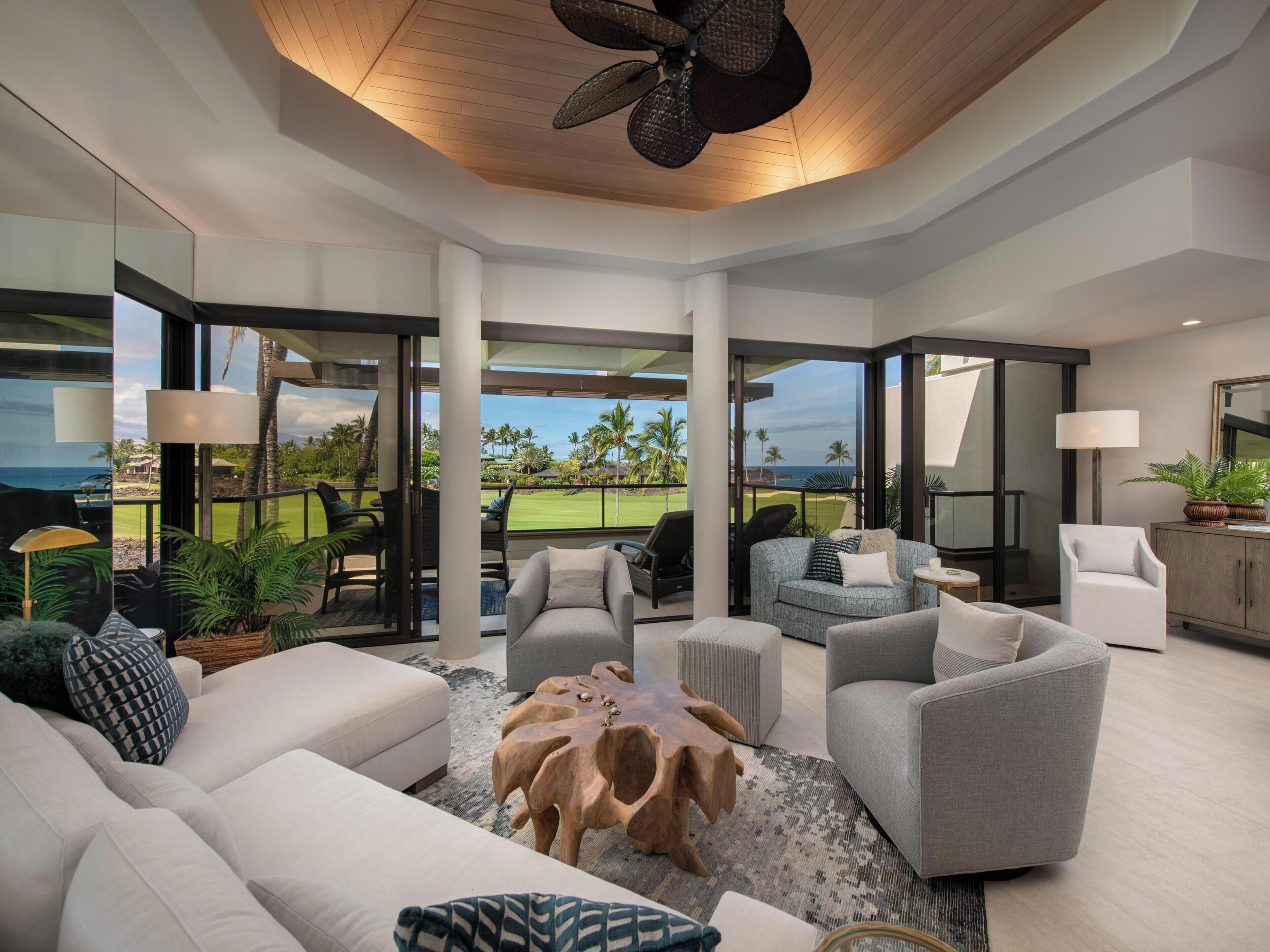 Un salon moderne avec vue sur un terrain de golf et l’océan. Il comprend un canapé, des fauteuils, une table basse et une lampe.