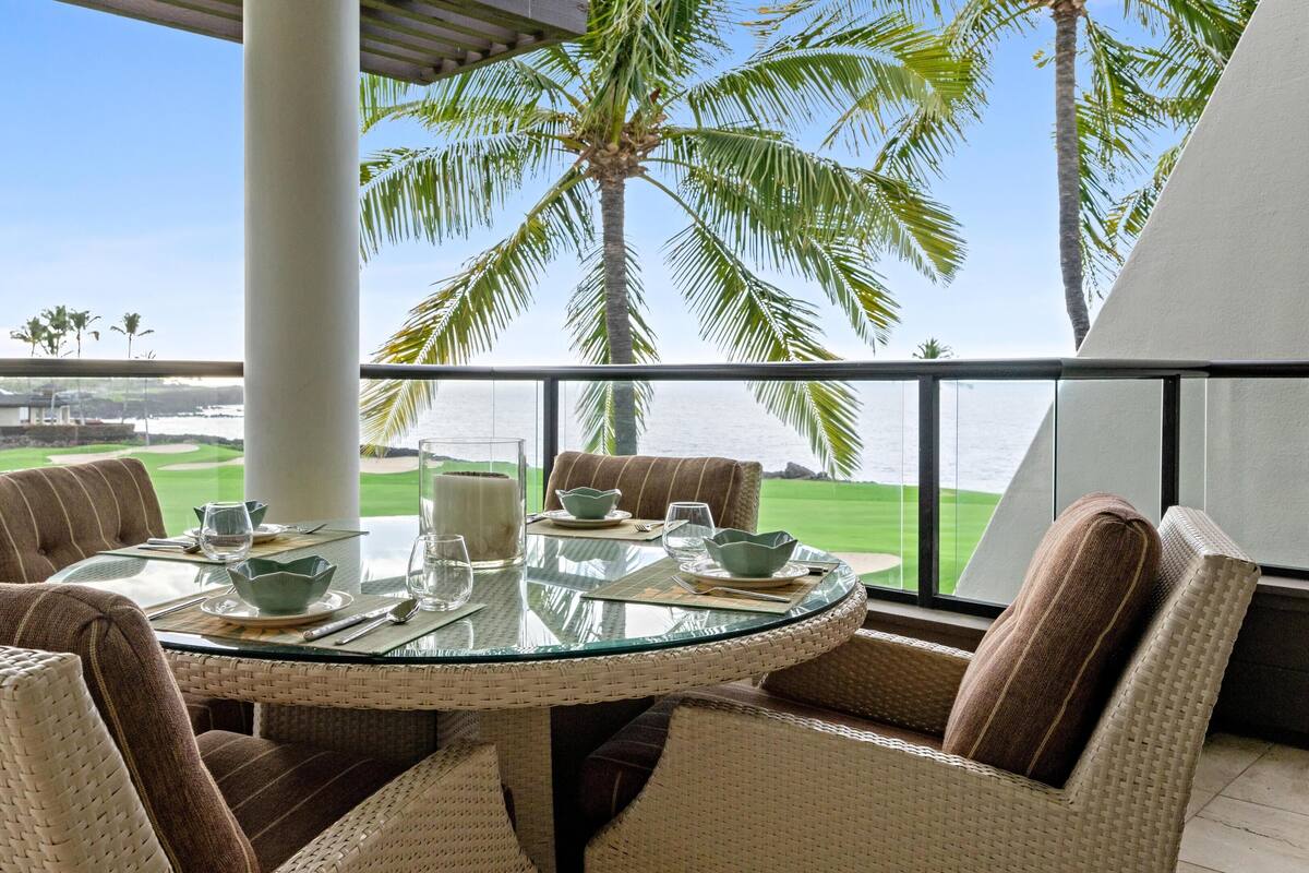 A round glass table with wicker chairs and a view of a golf course and ocean.