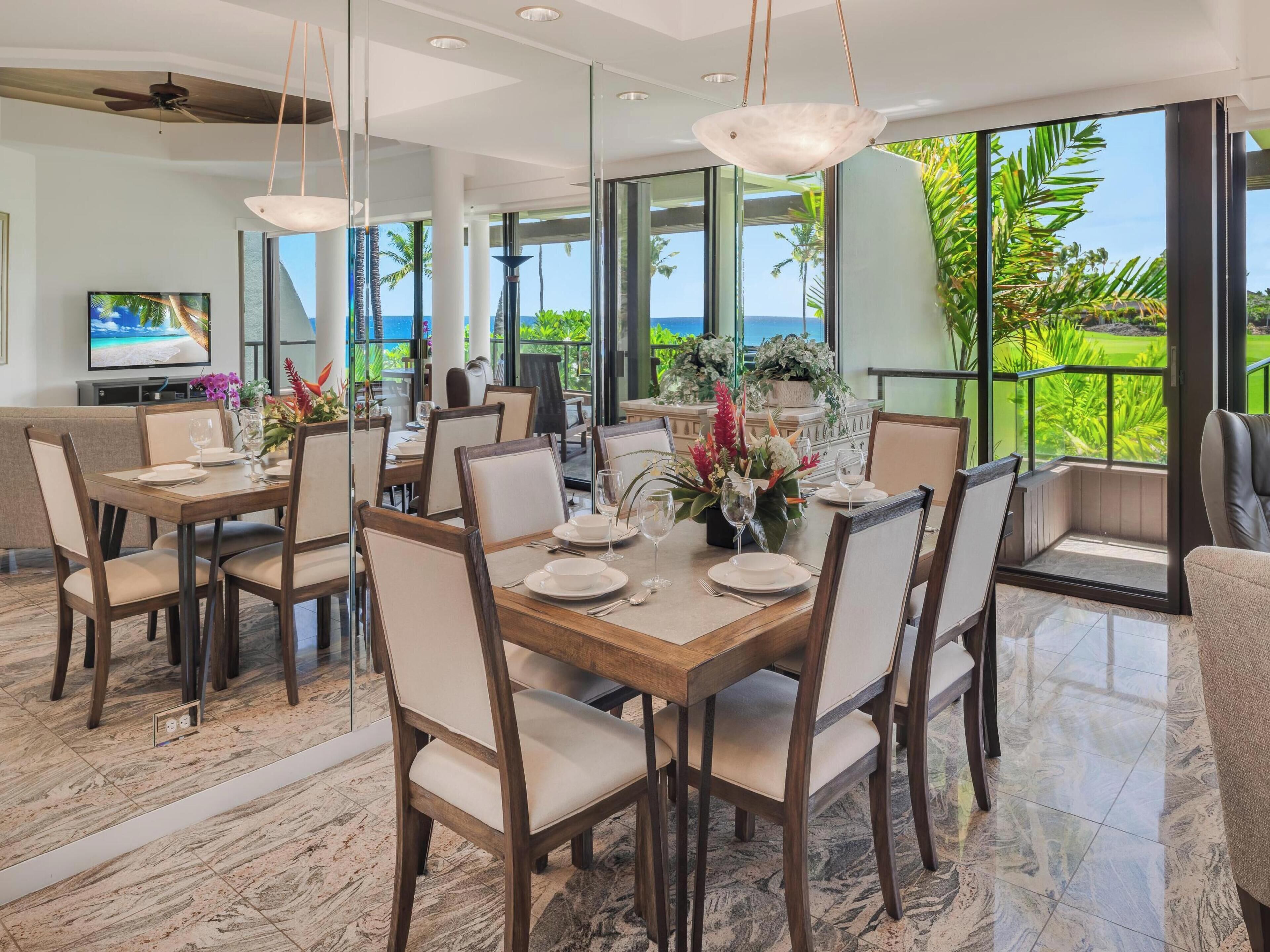 A modern dining area with a large table set for six, surrounded by chairs. The room has large windows offering a view of the ocean and palm trees.