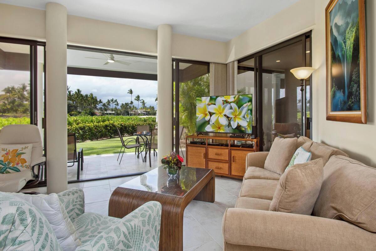 A spacious living room with a large flat-screen TV, comfortable seating, and a view of a lush garden.