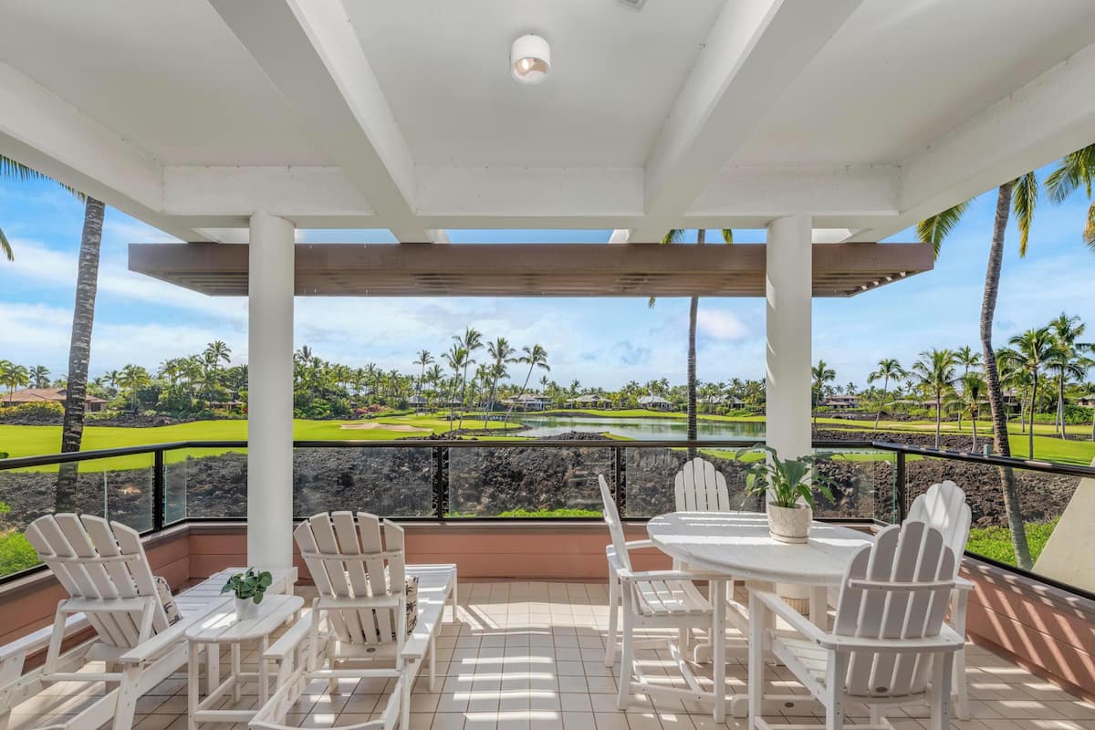 A covered outdoor seating area with white chairs and a table, overlooking a golf course and a body of water.