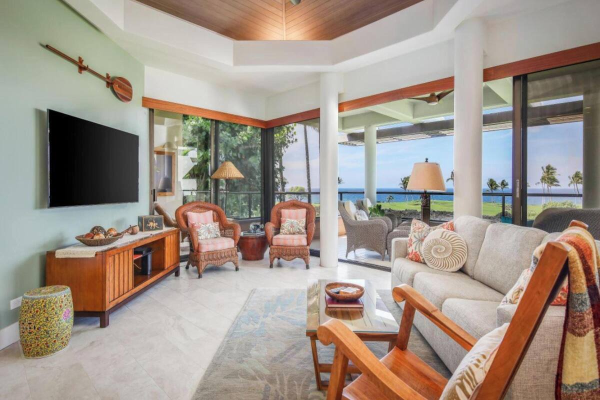 A spacious living room with a large TV, comfortable seating, and a view of the ocean.