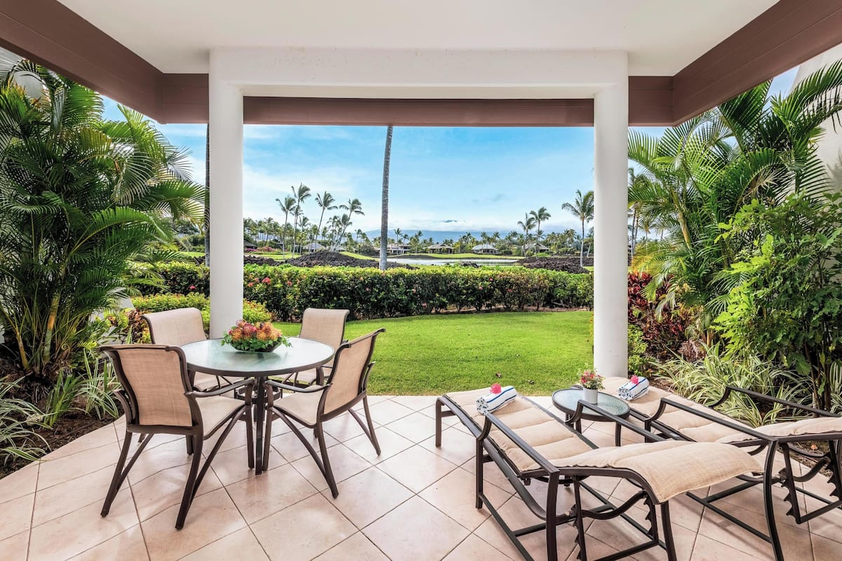 A covered patio with a round table and chairs, a lounge area with a sofa and armchairs, and a view of a lush garden and distant buildings.