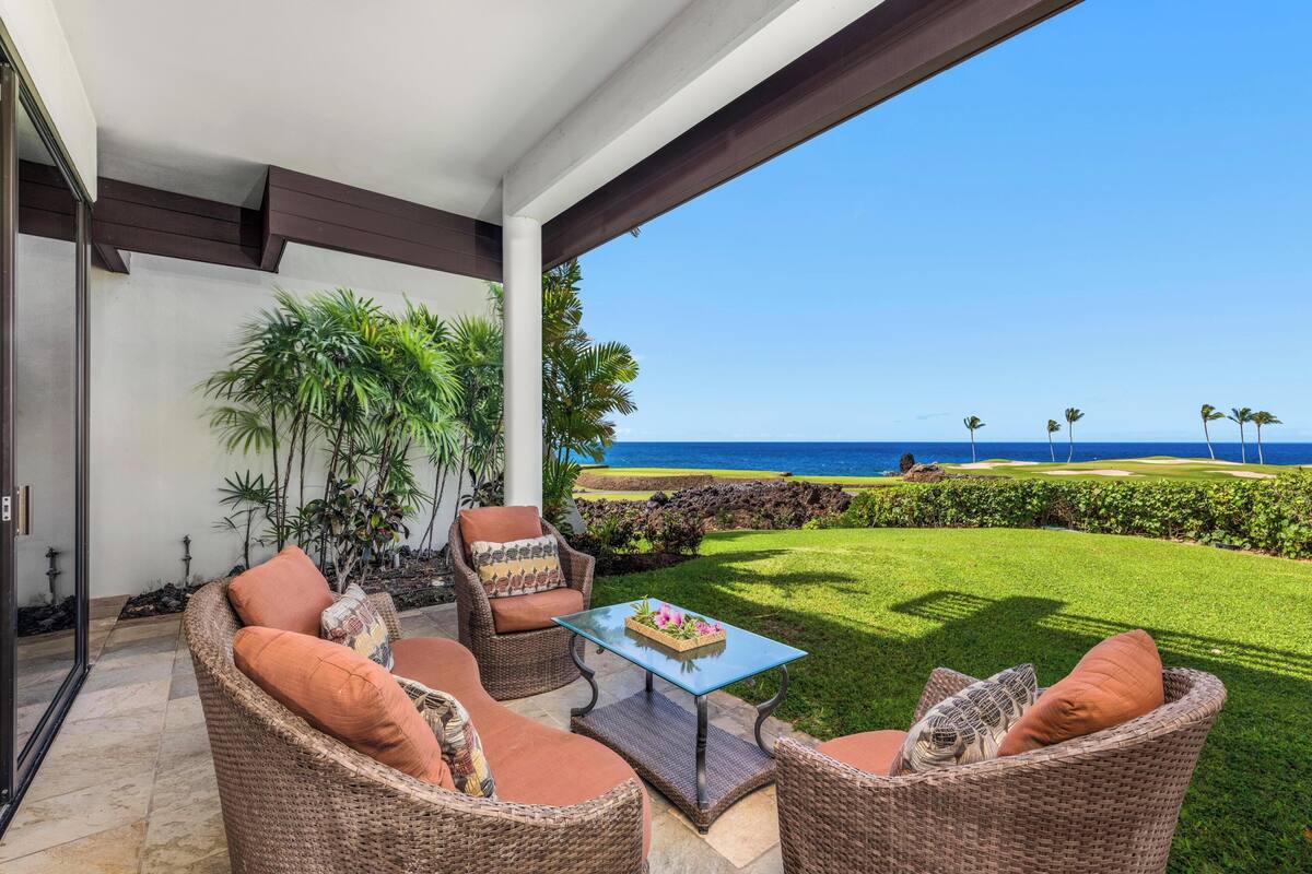 A covered outdoor seating area with wicker furniture and a glass table, overlooking a well-maintained lawn and ocean view.