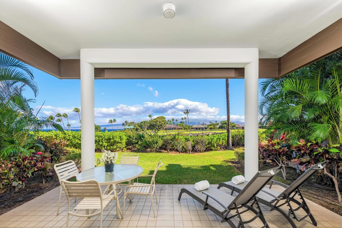 A covered patio with a table and chairs, a view of a lush garden and the ocean.
