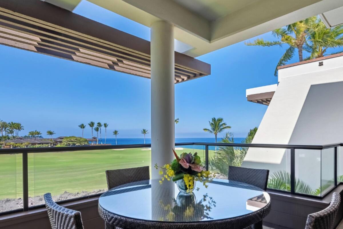 A balcony with a glass table and chairs, overlooking a golf course and the ocean.