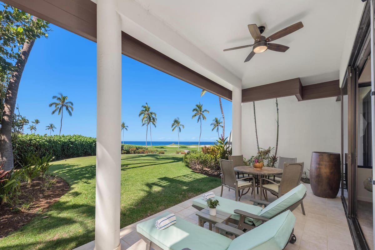 A covered outdoor area with a view of the ocean, a patio with lounge chairs, a table, and a ceiling fan.