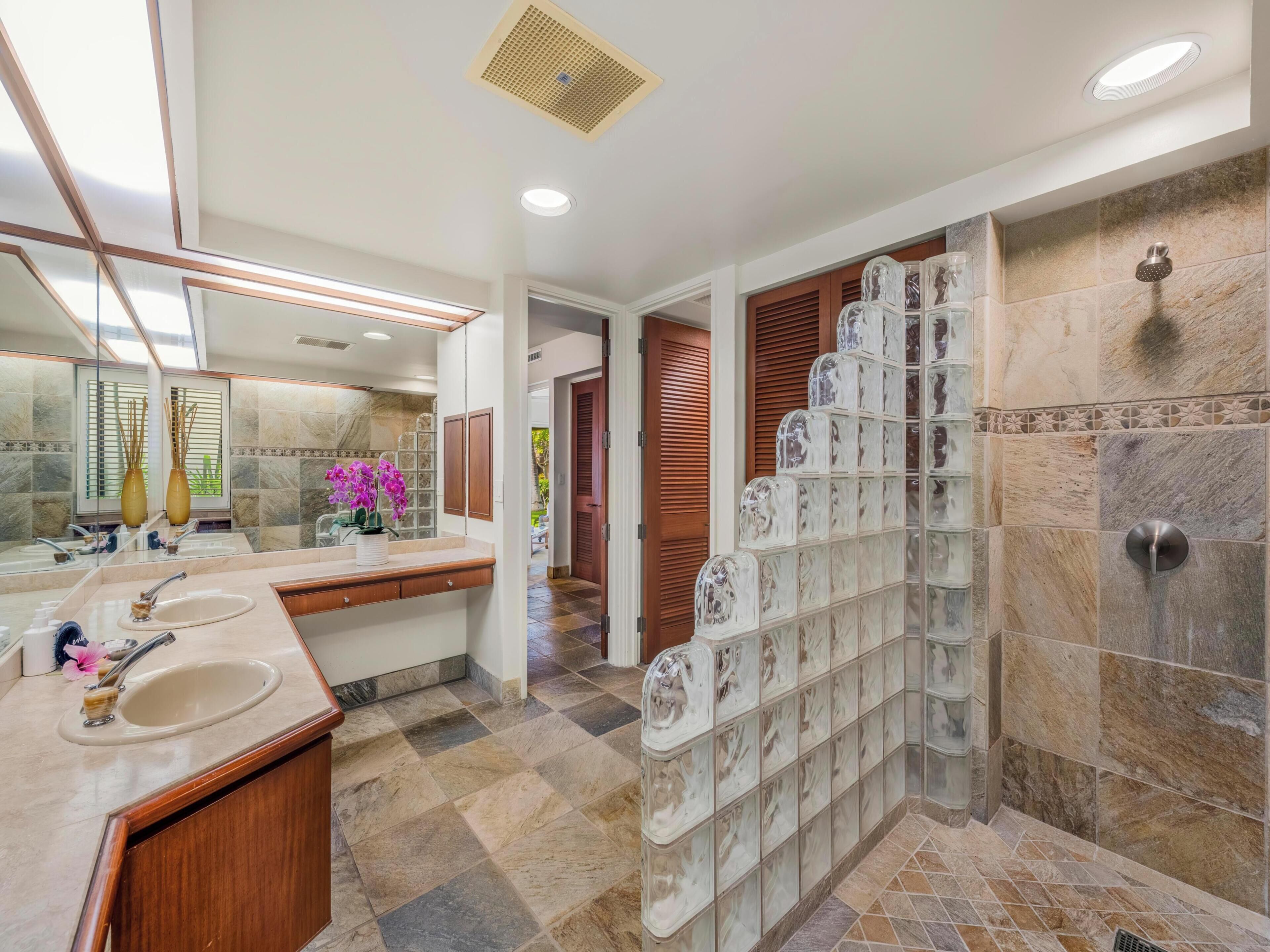 Un baño moderno con un espejo grande, dos lavamanos, una ducha con bloques de vidrio y piso de baldosas.