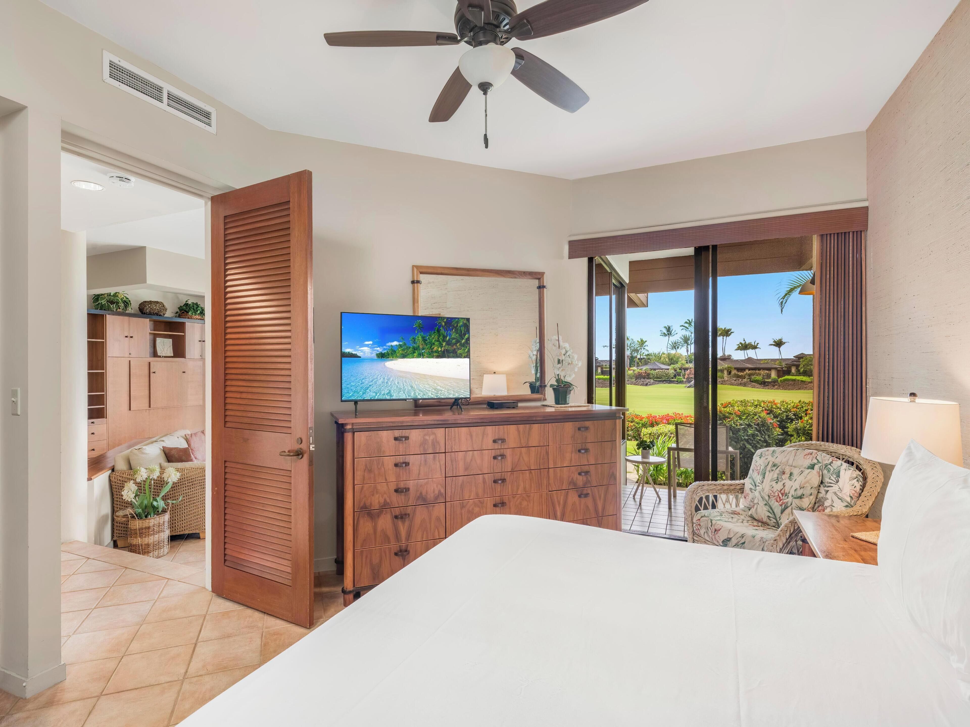 A bedroom with a bed, a TV on a wooden dresser, a ceiling fan, and a view of a golf course through large windows.