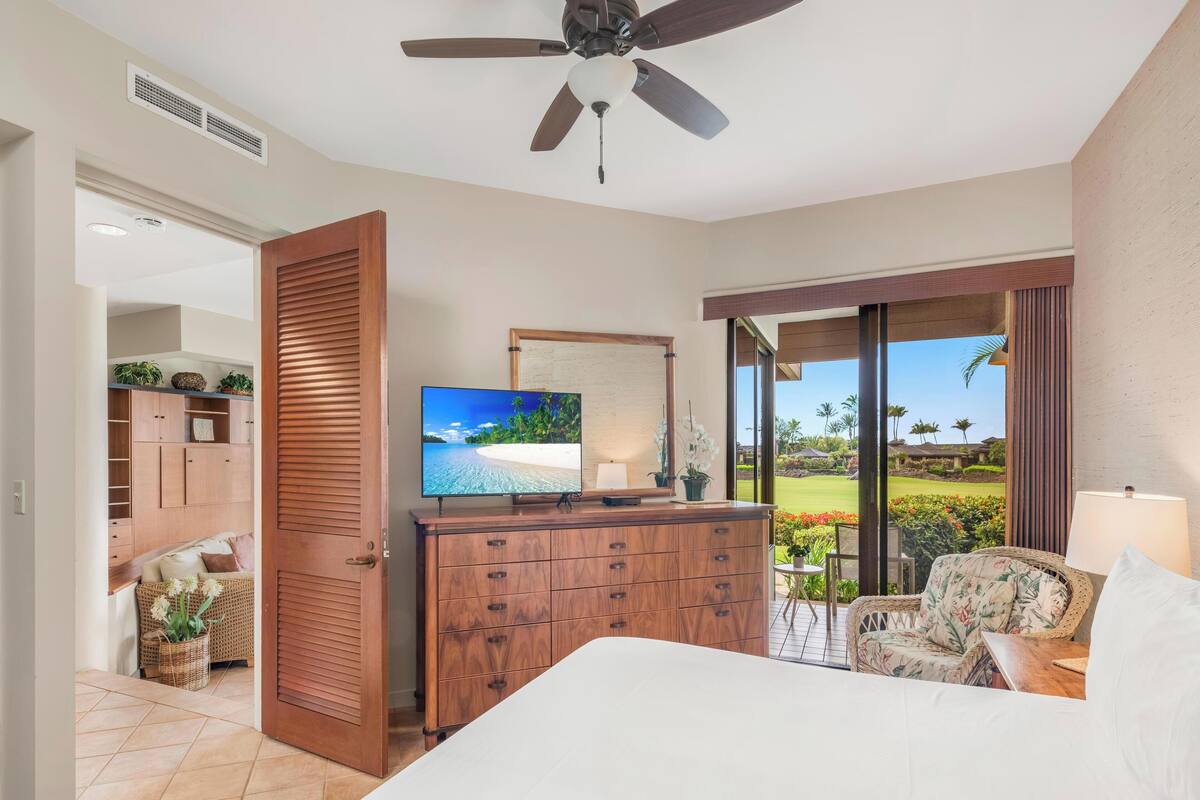A bedroom with a bed, a TV on a wooden dresser, a ceiling fan, and a view of a golf course through large sliding glass doors.