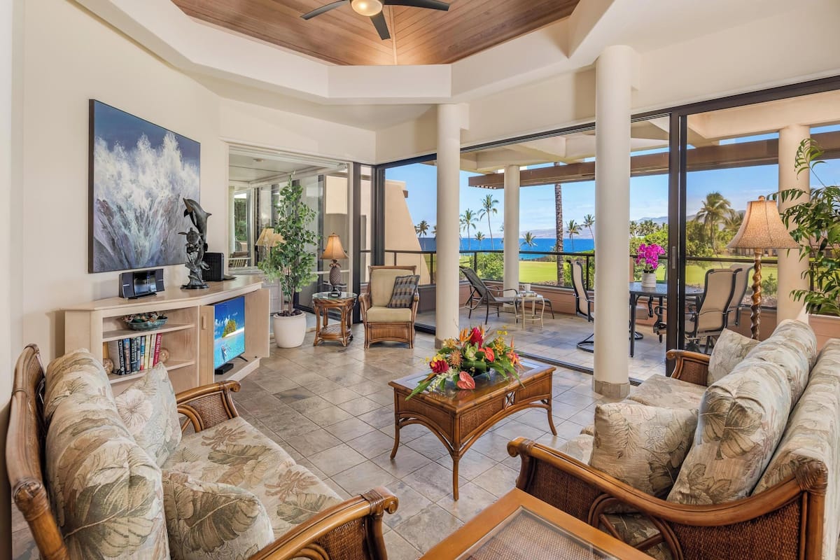 A spacious living area with a ceiling fan, comfortable seating, a television, and a view of the ocean.