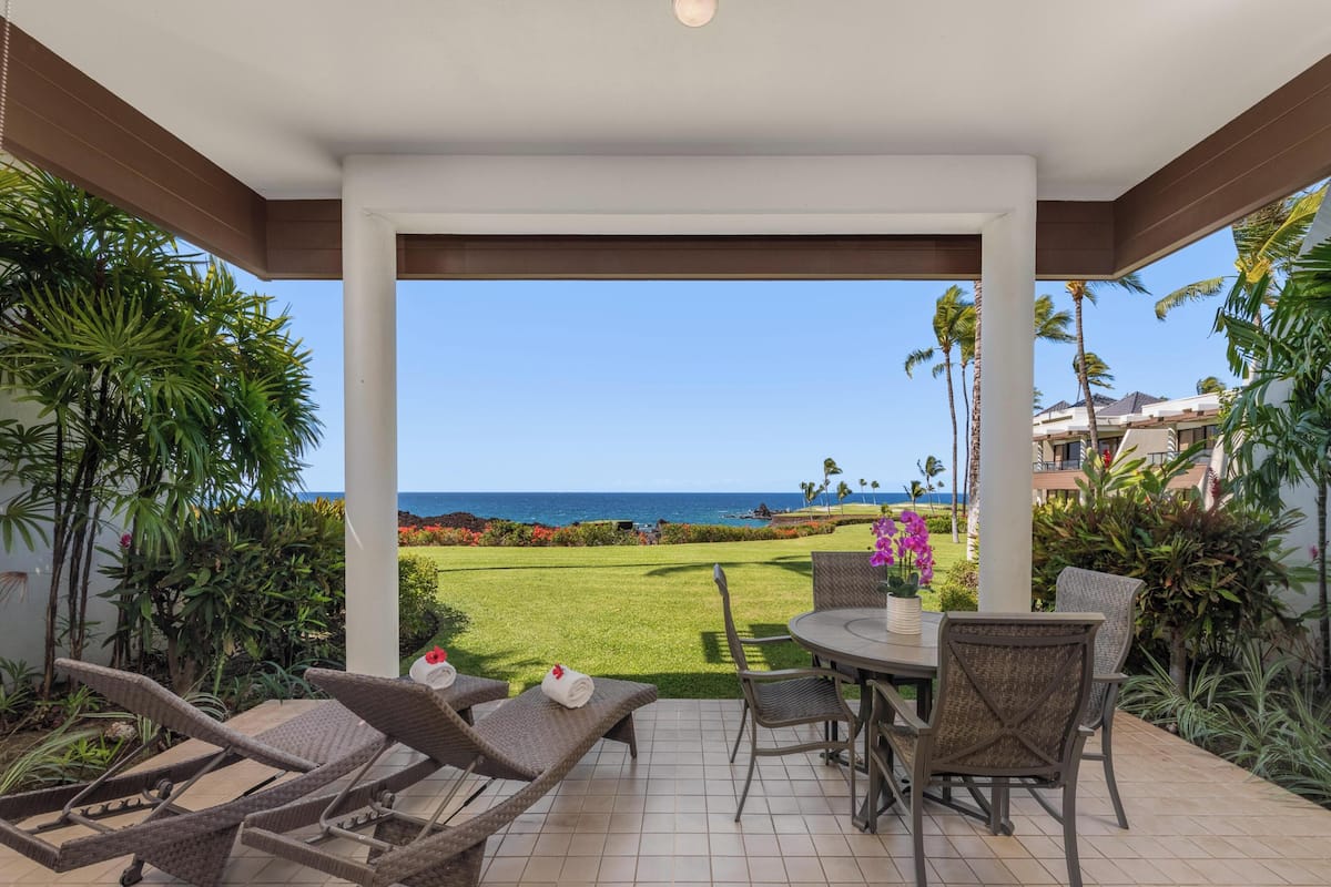 A covered patio with wicker furniture, a table with chairs, and a view of the ocean.