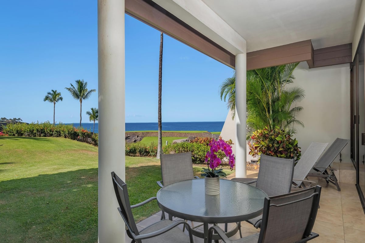 A covered outdoor seating area with a view of the ocean, featuring a round table and chairs, palm trees, and a flower arrangement.