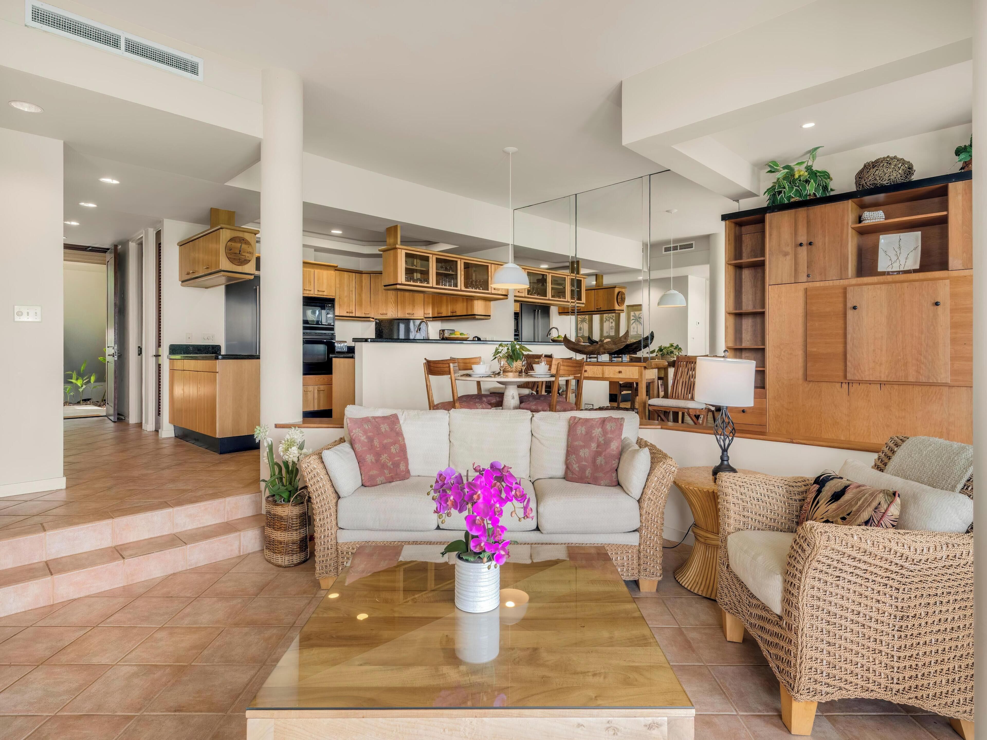 Amplia sala de estar con un conjunto de sofás, una mesa de centro y una mesa de comedor con sillas. Se observa una cocina al fondo con armarios de madera y un diseño moderno.