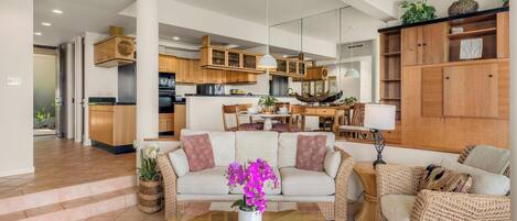 Amplia sala de estar con un conjunto de sofás, una mesa de centro y una mesa de comedor con sillas. Se observa una cocina al fondo con armarios de madera y un diseño moderno.