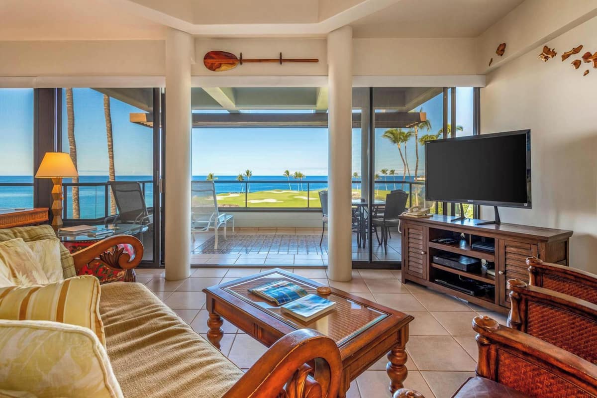 A spacious living room with a large TV, comfortable seating, a coffee table, and a view of the ocean.