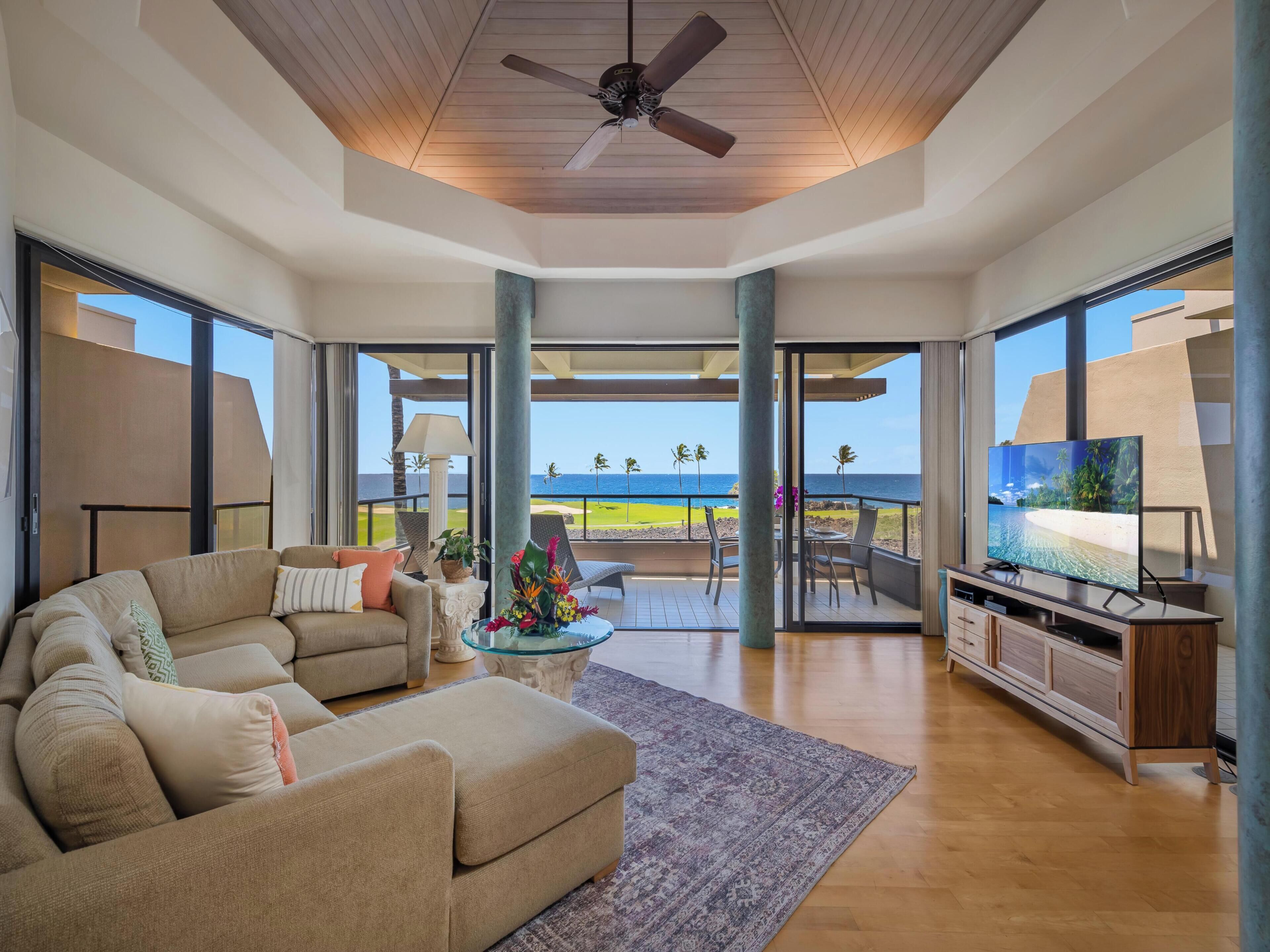 A spacious living room with a large beige sofa, a coffee table with a floral centerpiece, and a television on a wooden cabinet. The room has large glass doors leading to a balcony with a view of the ocean.