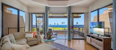 A spacious living room with a large beige sofa, a coffee table with a floral centerpiece, and a television on a wooden cabinet. The room has large glass doors leading to a balcony with a view of the ocean.