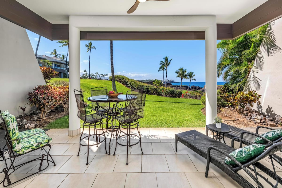 A covered patio with a table and chairs, a ceiling fan, and a view of a grassy area with palm trees and a body of water.