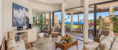 A spacious living area with a ceiling fan, a large painting, and a view of the ocean through large windows.