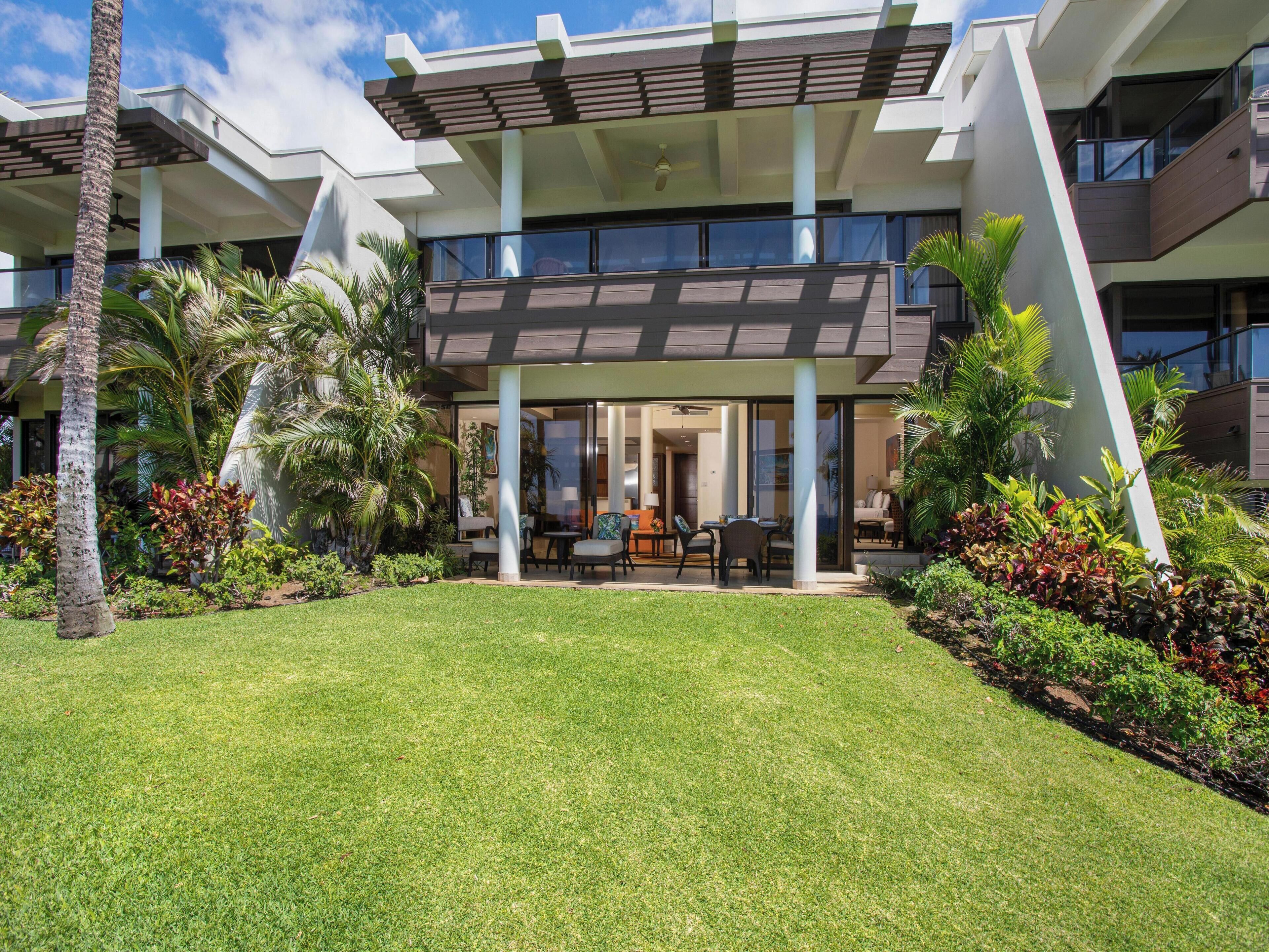 A modern building with a covered outdoor seating area, surrounded by lush greenery and palm trees.