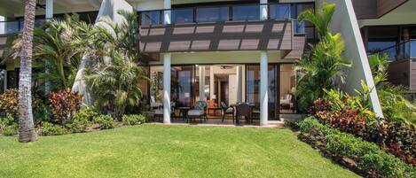 A modern building with a covered outdoor seating area, surrounded by lush greenery and palm trees.