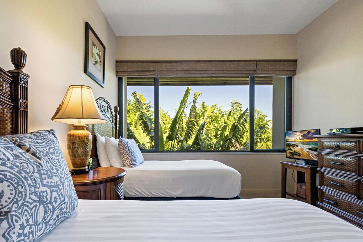 A hotel room with two beds, a lamp, a TV, and a window with a view of greenery.