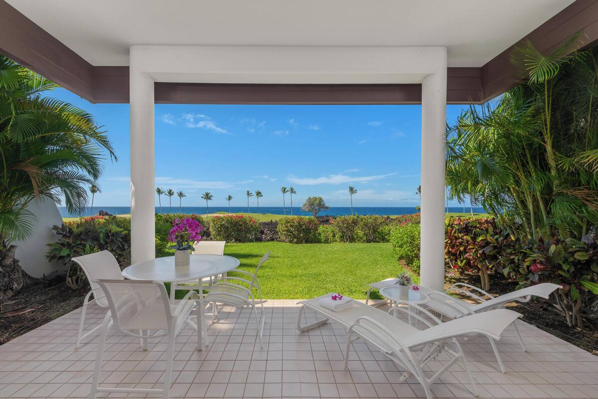 A covered patio with a view of the ocean, white chairs, and a table with a vase of flowers.