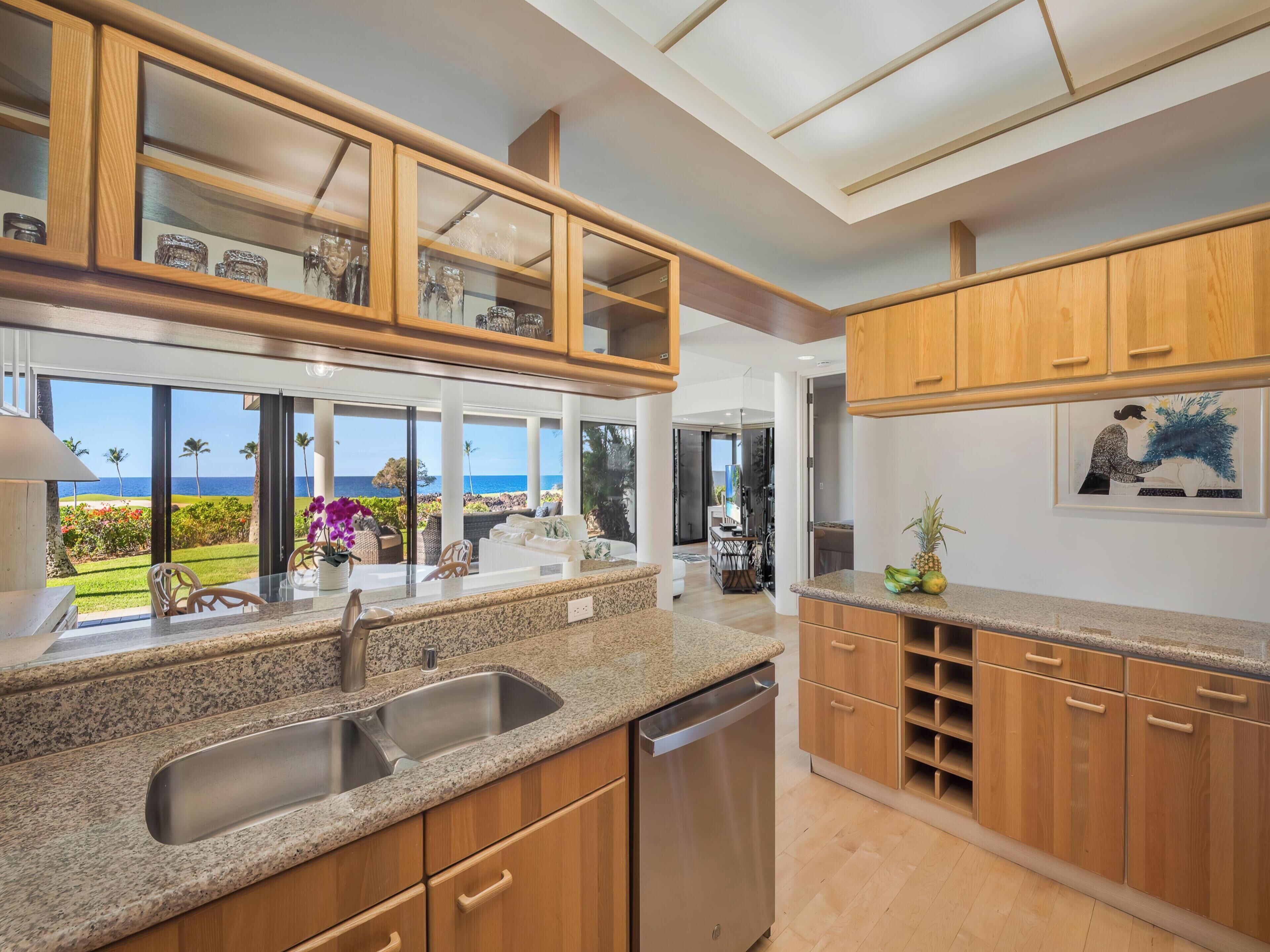 A modern kitchen with granite countertops, wooden cabinets, and a large window overlooking a garden and ocean view.