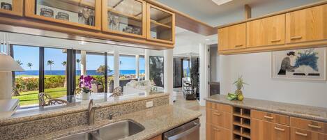 A modern kitchen with granite countertops, wooden cabinets, and a large window overlooking a garden and ocean view.