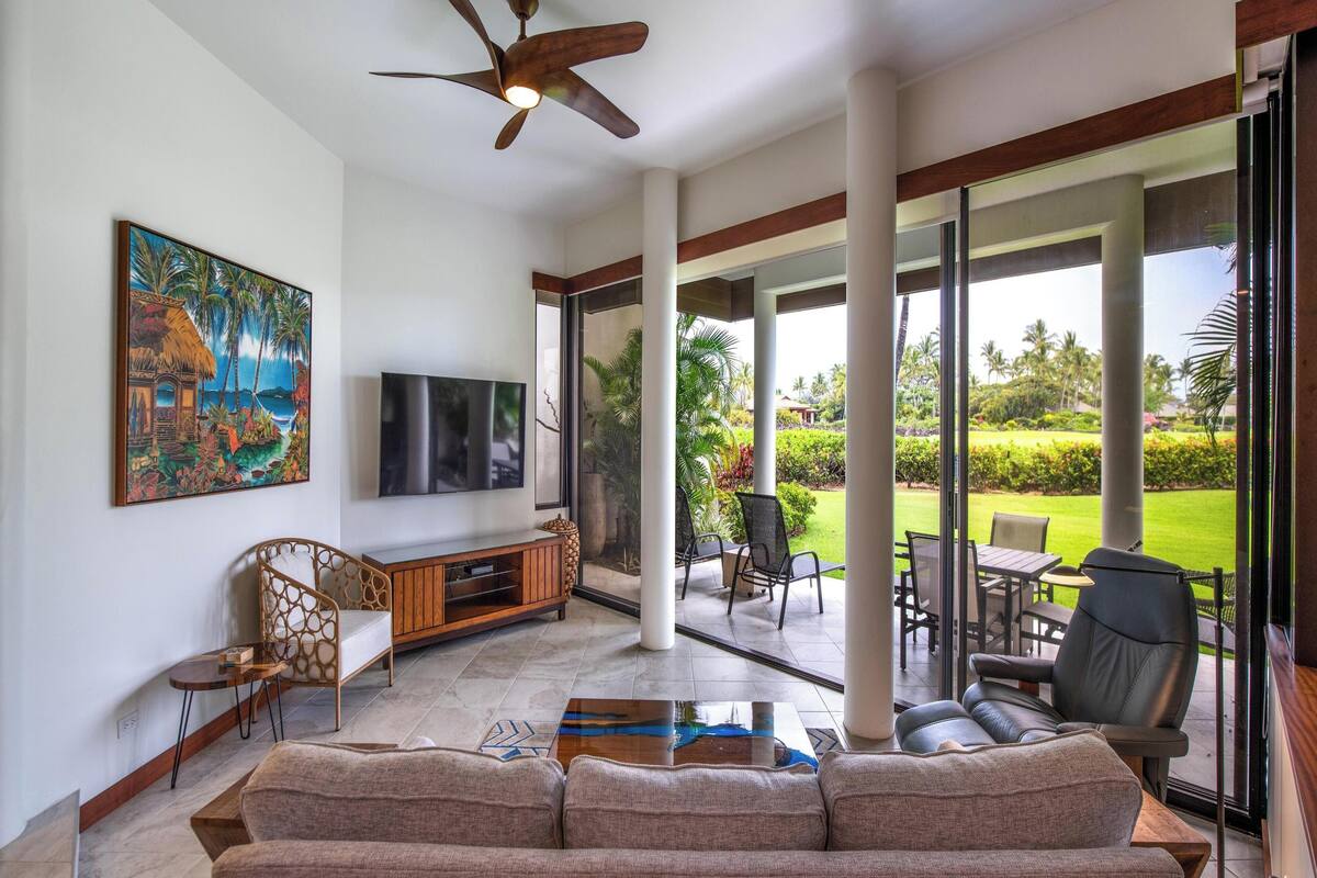 A spacious living room with a large flat-screen TV, a ceiling fan, and a large window offering a view of a lush garden.