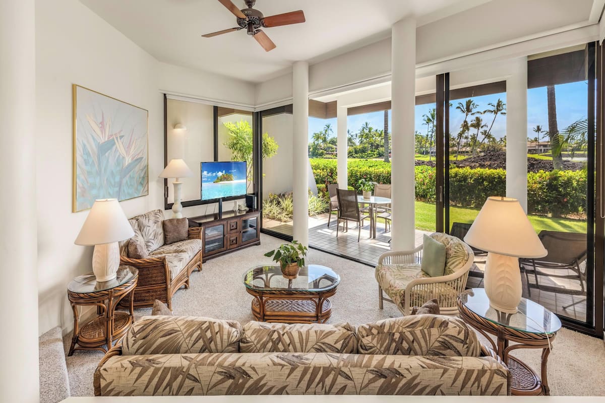 A spacious living area with a television, wicker furniture, and large windows offering a view of a landscaped garden.