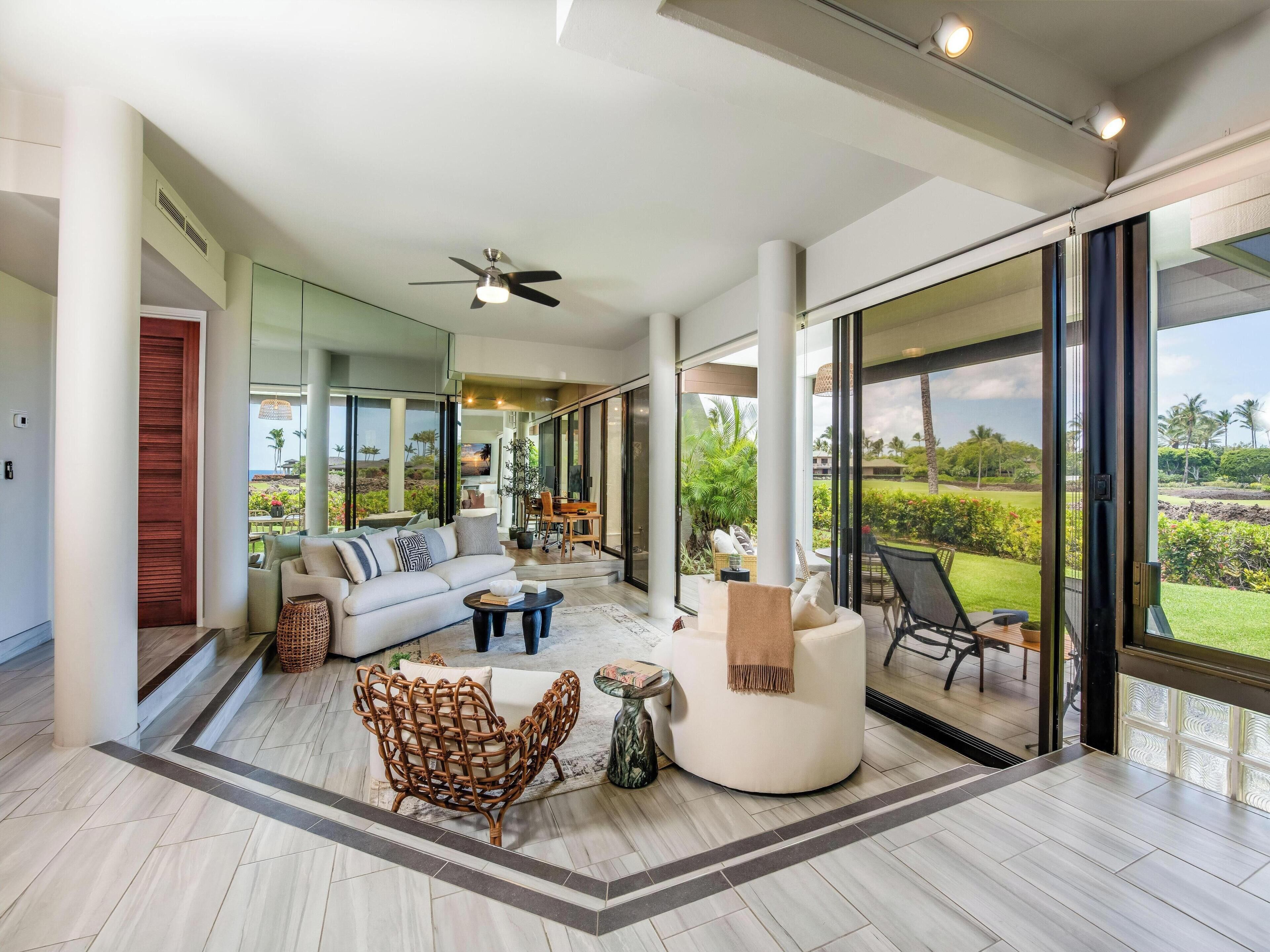 A spacious living room with a white sofa, a rattan chair, and a coffee table. There is a ceiling fan and large windows offering a view of the outdoors.