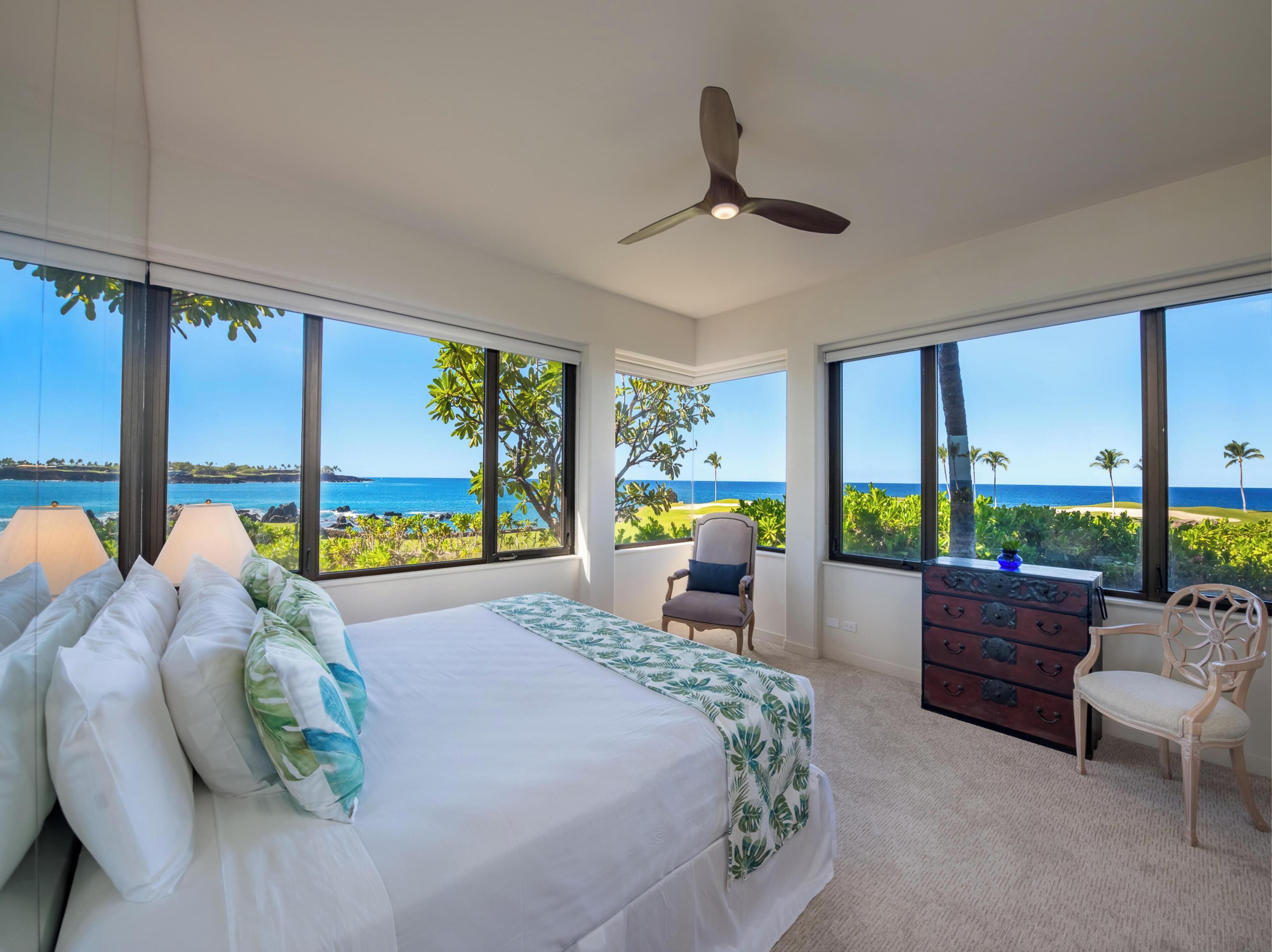 A bedroom with a large bed, a dresser, a chair, and a ceiling fan. It features large windows with a view of the ocean and palm trees.