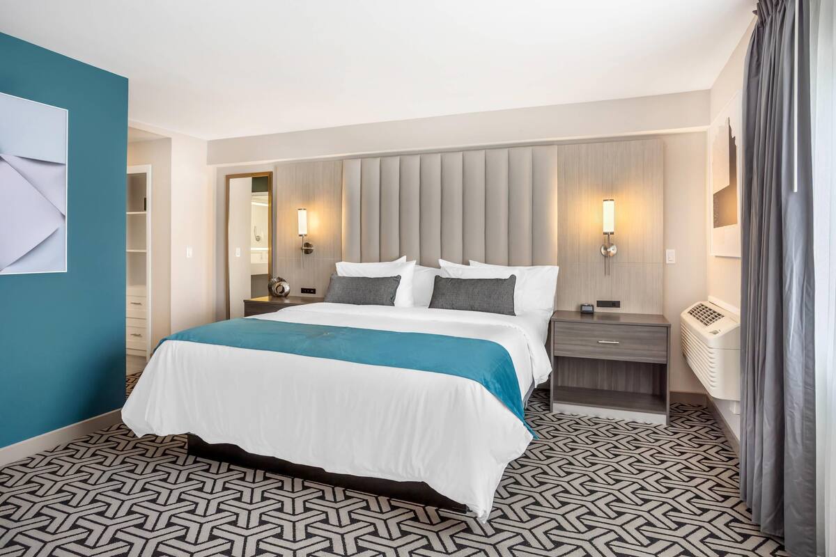 Una habitación de hotel con una cama grande, cabecera con paneles verticales, lámparas de noche, mesitas de noche, calentador incorporado y piso con diseño.