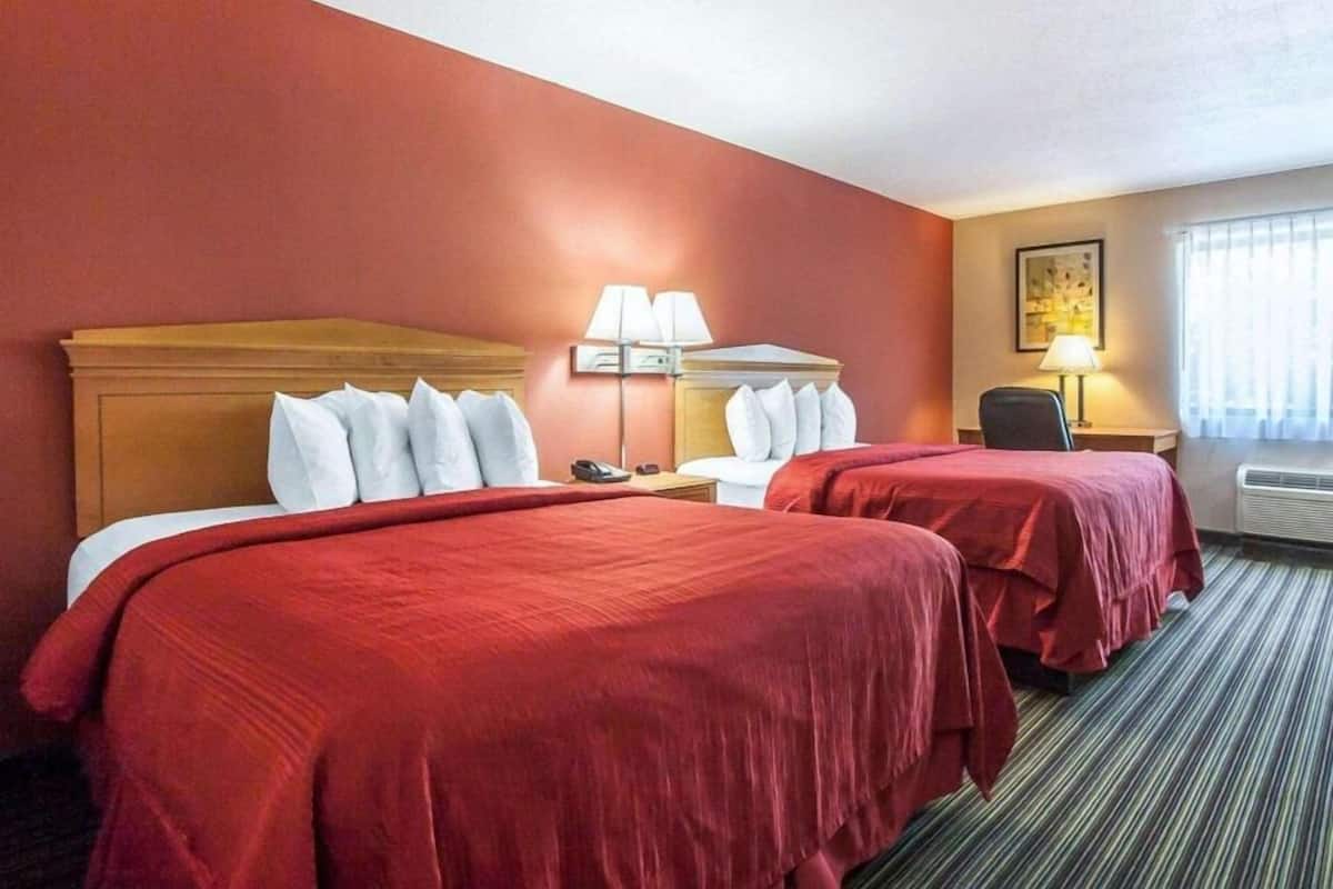 Habitación de hotel con dos camas, cabeceros, mesitas de noche, lámparas, una silla y un ventanal con cortinas.