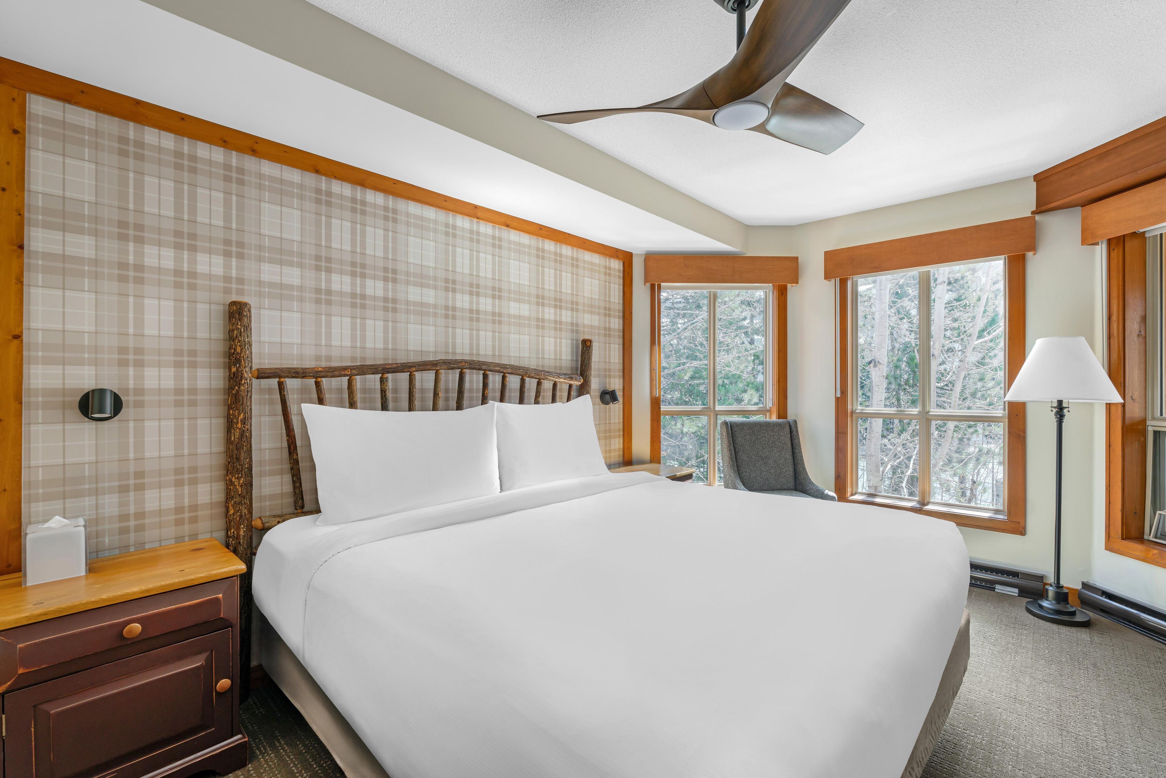 Une chambre d’hôtel avec un grand lit, une tête de lit en bois, une table de chevet, une lampe et une fenêtre donnant sur des arbres.