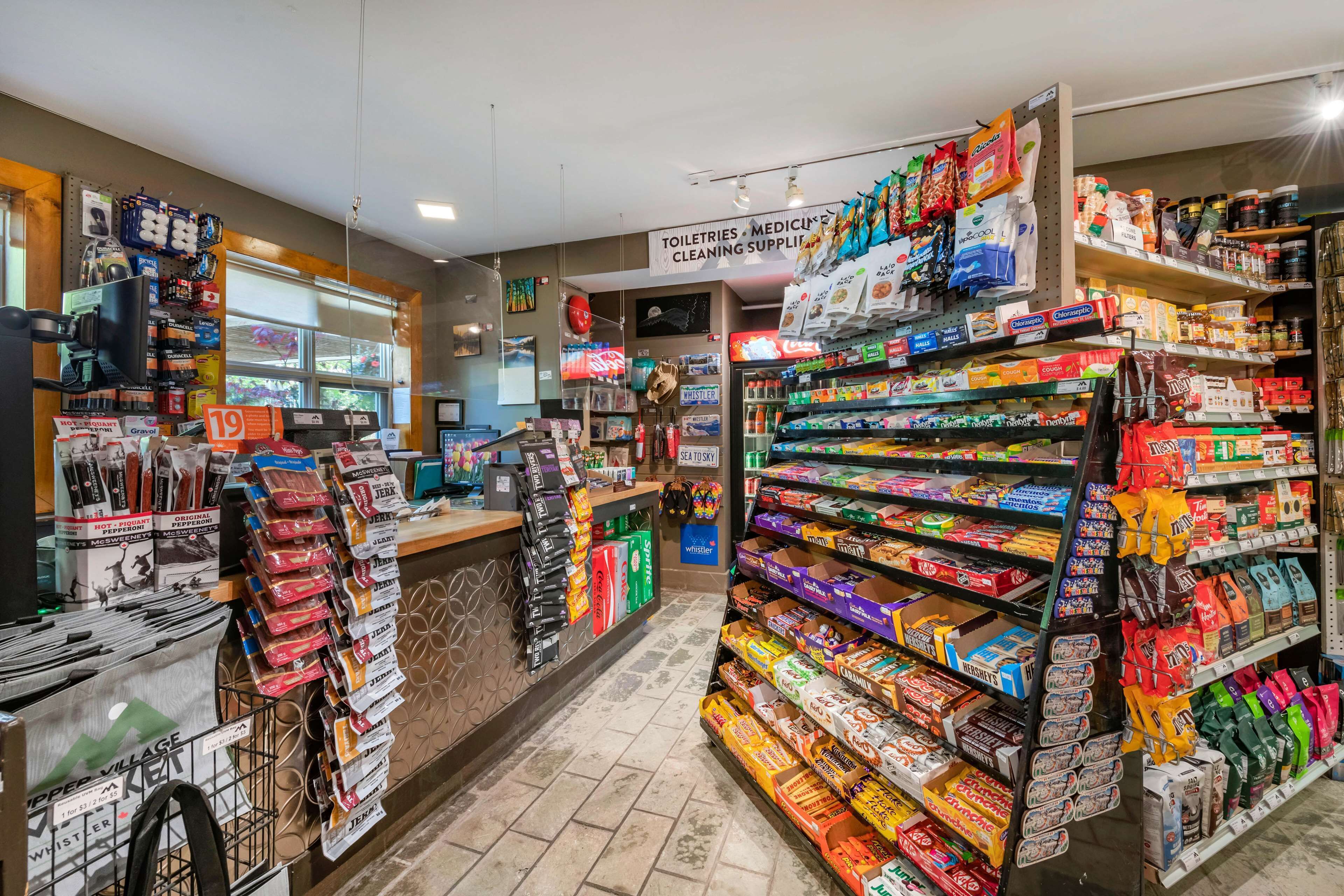 L’intérieur d’une épicerie, avec des étagères remplies de divers produits, un comptoir de caisse et un panneau indiquant la vente d’articles de toilette et de produits d’entretien.