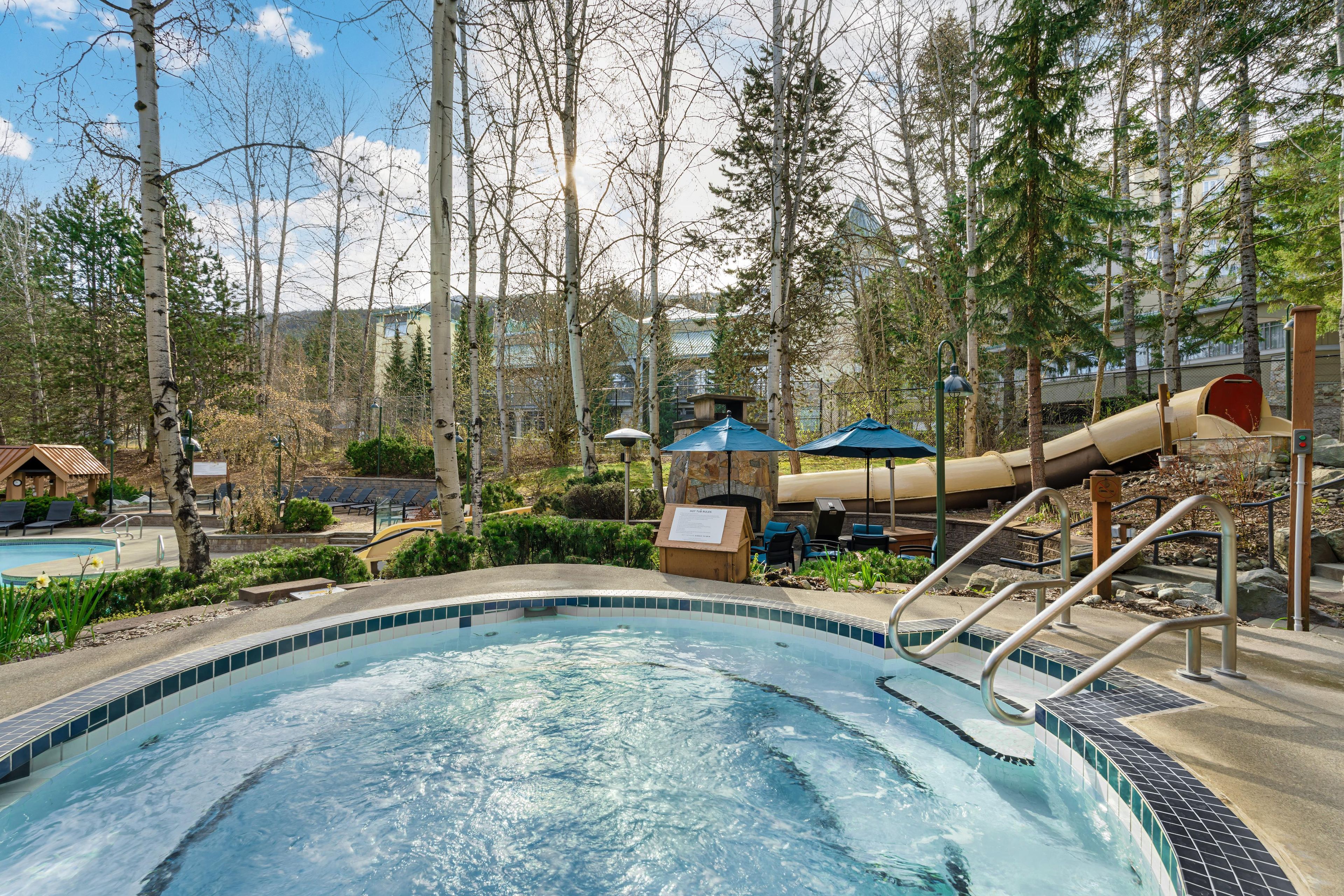 Une piscine avec un toboggan, entourée d’arbres et dotée d’un espace de détente.