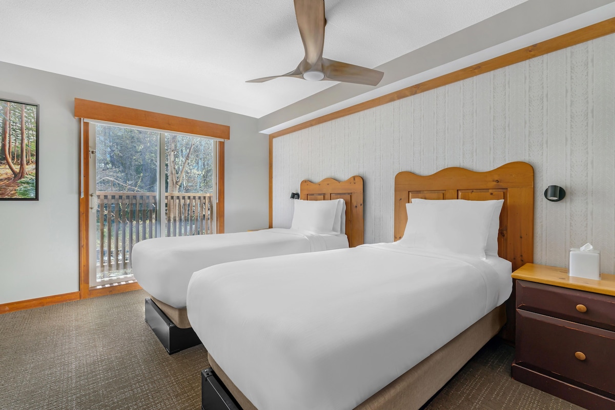 Une chambre d’hôtel avec deux lits, un ventilateur de plafond, une tête de lit en bois, une table de chevet et une vue sur l’extérieur grâce à une porte coulissante.
